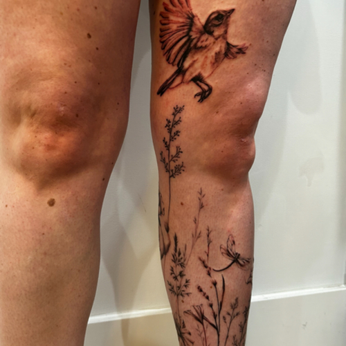 A delicate fine line wildflower wrap-around tattoo that gracefully encircles the leg. This design features intricate, detailed blossoms and stems, creating a natural and elegant look. 