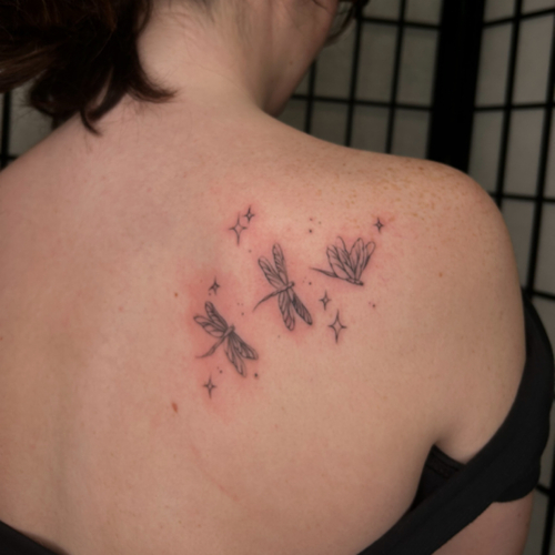 A fine line dragonfly tattoo elegantly placed on the shoulder blade, featuring delicate and intricate details. The design showcases the grace and beauty of dragonflies, with their slender bodies and delicate wings rendered in a minimalist style!